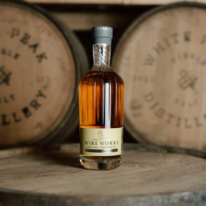 A bottle of Wire Works Calvados Arrivee english whisky stands on a wooden barrel with two blurred White Peak Distillery barrels in the background.