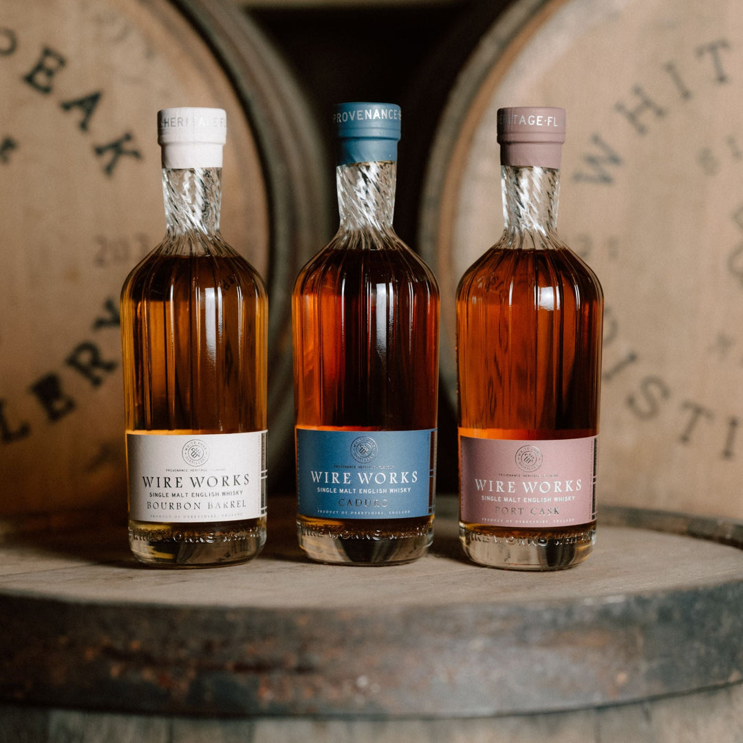 Three bottles of Wire Works whiskey with different colored labels on a wooden barrel background.