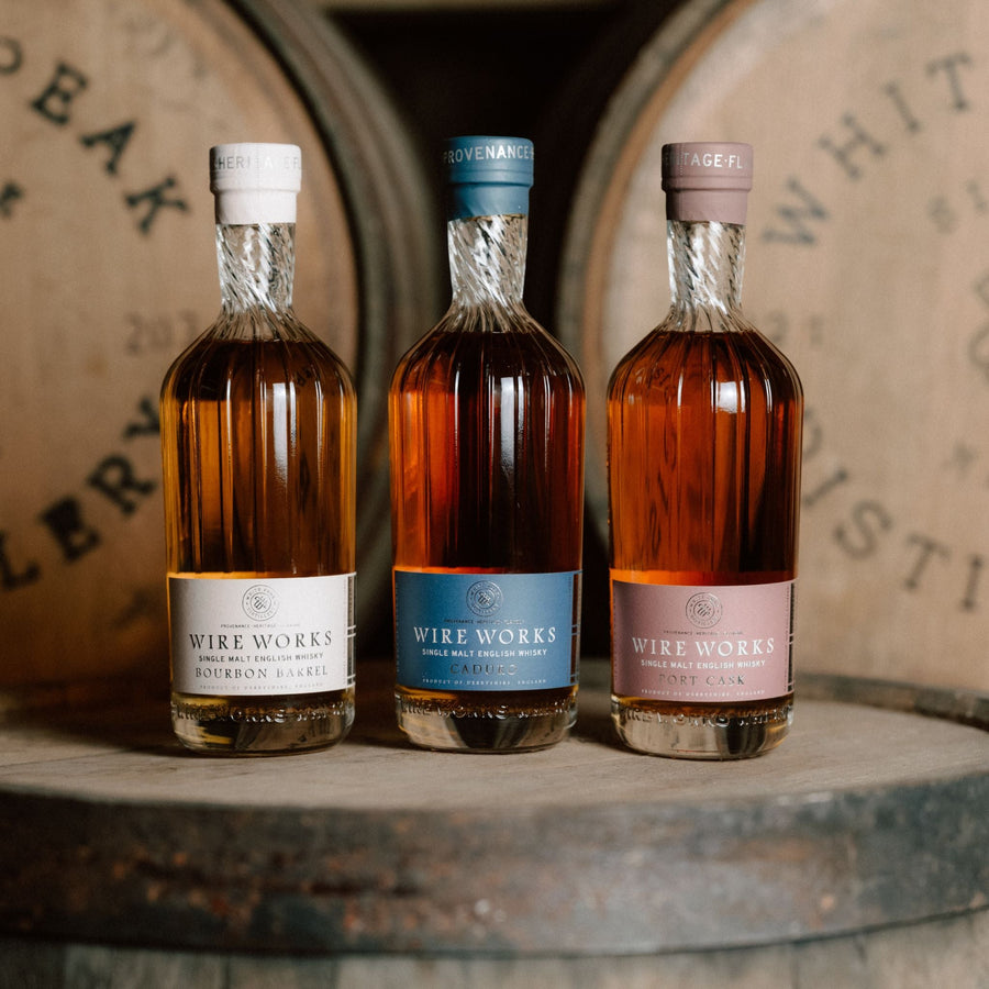 Three bottles of Wire Works whiskey with different colored labels on a wooden barrel background.