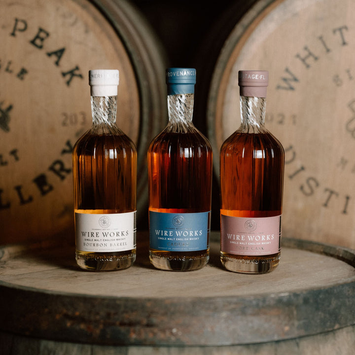 Three bottles of Wire Works whiskey in front of wooden barrels.
