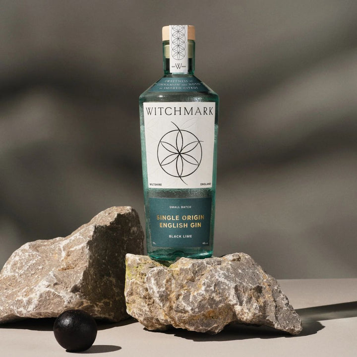 A bottle of Witchmark Black Lime Single Origin English Gin with a clear label on a rock with a black lime