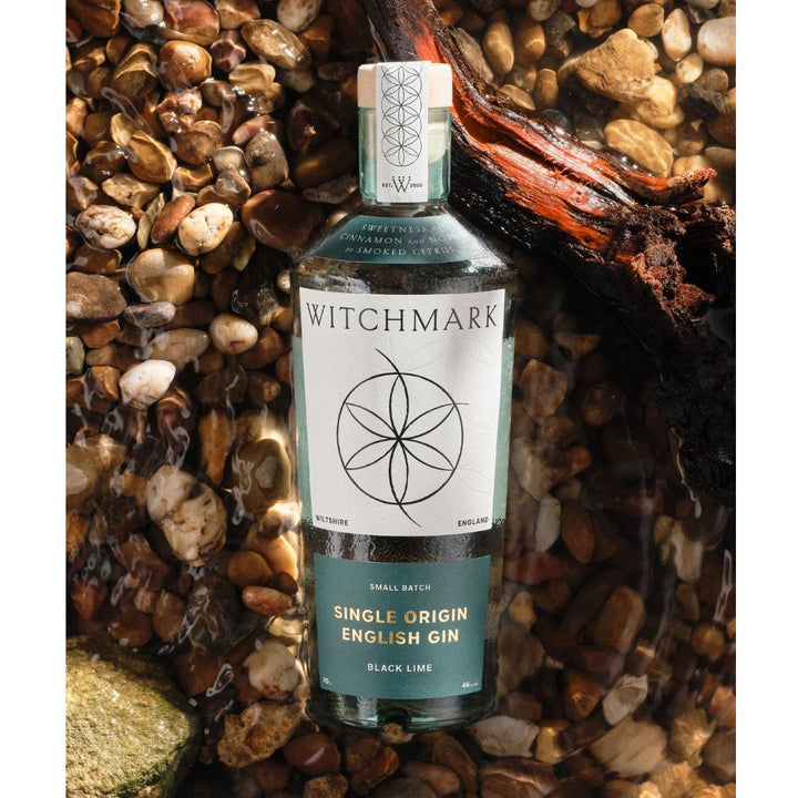 A bottle of Witchmark Black Lime Single Origin English Gin with a clear label and a witchmark design on the front resting on some pebbles in a stream