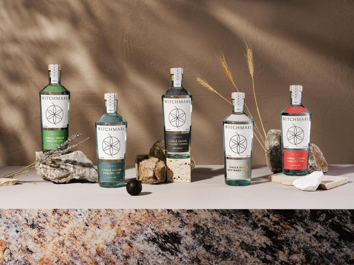 Five Witchmark spirit bottles in assorted green, blue-green, black, and red arranged on a stone surface with natural props like rocks, dried wheat, and lavender against a textured brown backdrop.