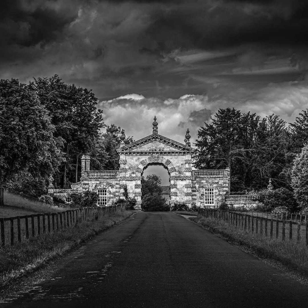 Black and white landscape photograph of a grand stone archway or gatehouse centered on a road, surrounded by dense trees under a dramatic cloudy sky.