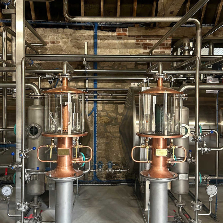 Industrial distillation apparatus with copper and stainless steel columns, glass collectors, and piping inside Witchmark distillery.