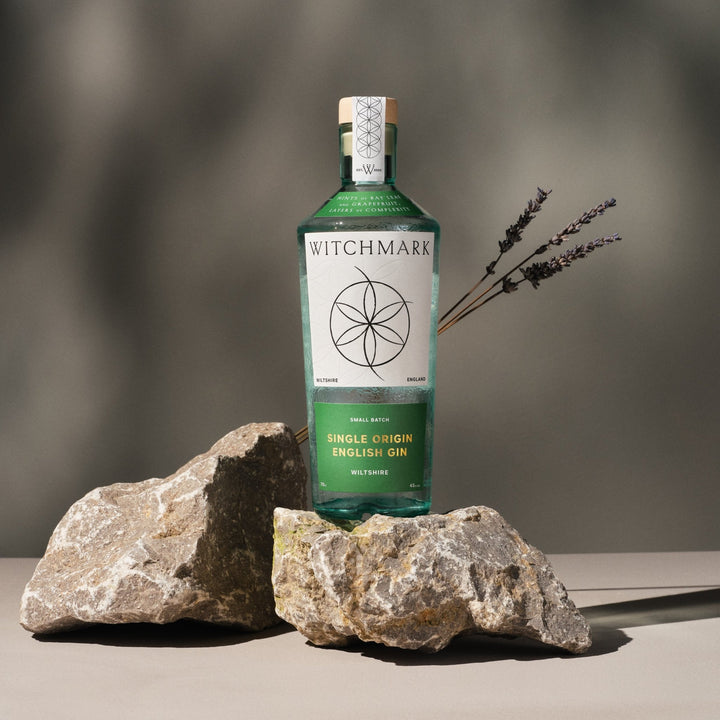 Bottle of Witchmark gin with a label featuring a witchmark logo, placed on a neutral background with rocks.