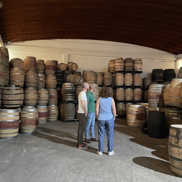 Three people standing in a barrel room surrounded by stacked wooden barrels under a dark wooden ceiling.