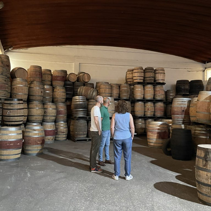 Three people standing in a barrel room surrounded by stacked wooden barrels under a dark wooden ceiling.